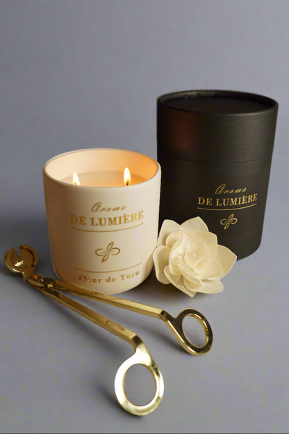 Fleur de Yuzu luxury candle by Aroma De Lumière in a matte white jar with gold foil branding, shown lit with a soft glow beside a black cylindrical gift box, cream flower accent, and gold candle care tools — a fresh and refined addition to any modern home