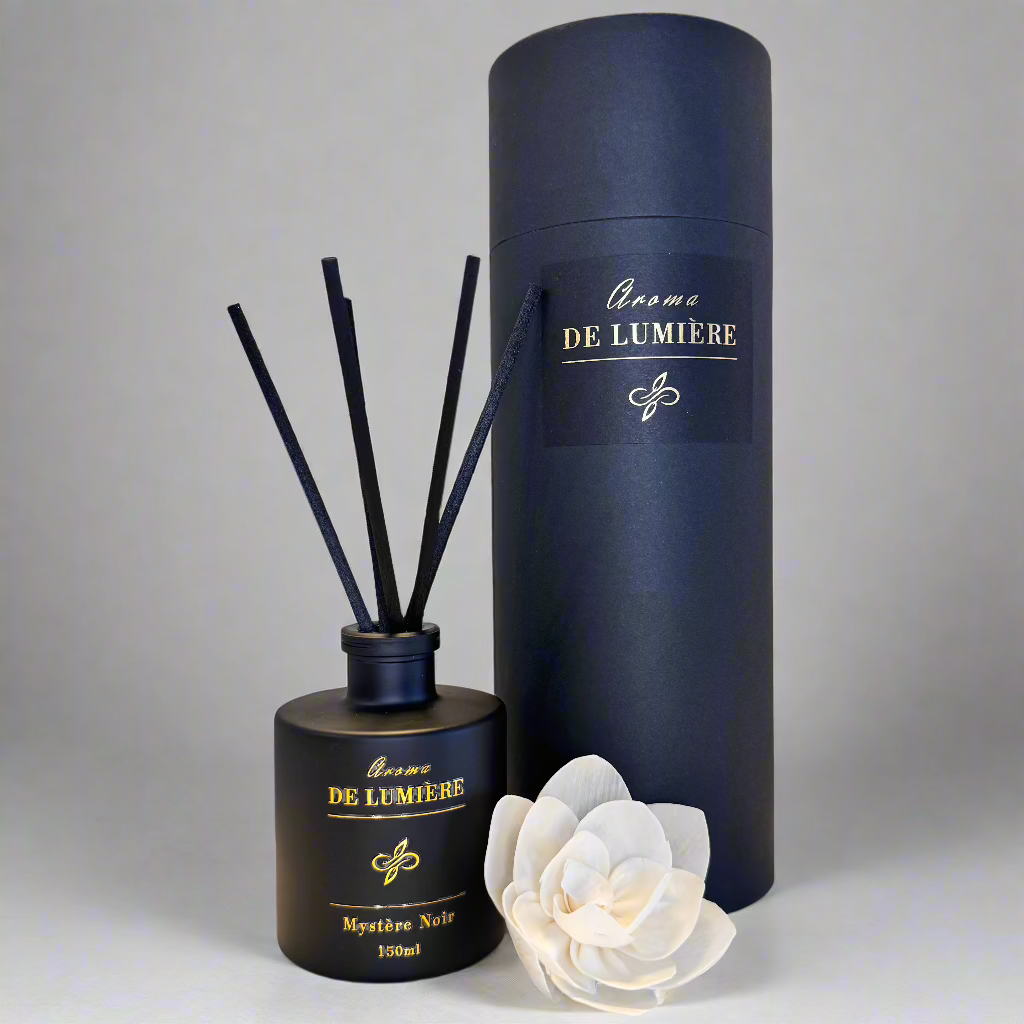 Mystère Noir reed diffuser by Aroma De Lumière in a matte black 150ml bottle with elegant gold foil branding, paired with a matching cylindrical gift box and soft cream flower accent — a luxurious home fragrance set crafted for a bold, modern aesthetic