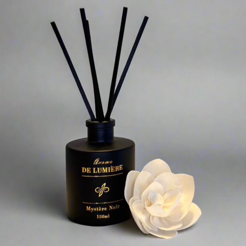 Mystère Noir luxury reed diffuser by Aroma De Lumière, showcased in a sleek matte black 150ml bottle with gold foil detailing, accompanied by elegant black reeds and a soft cream floral accent — designed to evoke a bold, mysterious ambiance in any space
